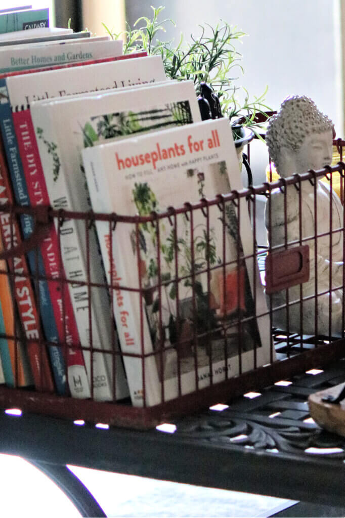 A metal basket with various books inside. Along with a small Buddha and a faux plant