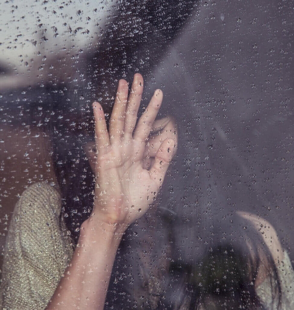 A woman stands on the inside while it rains outside. She has her hand on the window and she is in shadow, feeling caught in the headlights.