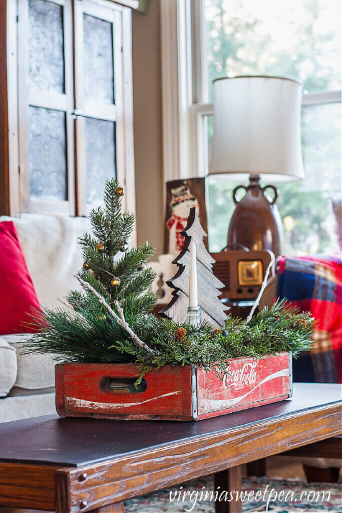 A vintage coco-cola box used as a container to hold Christmas greenery, a candle and a couple of small trees
