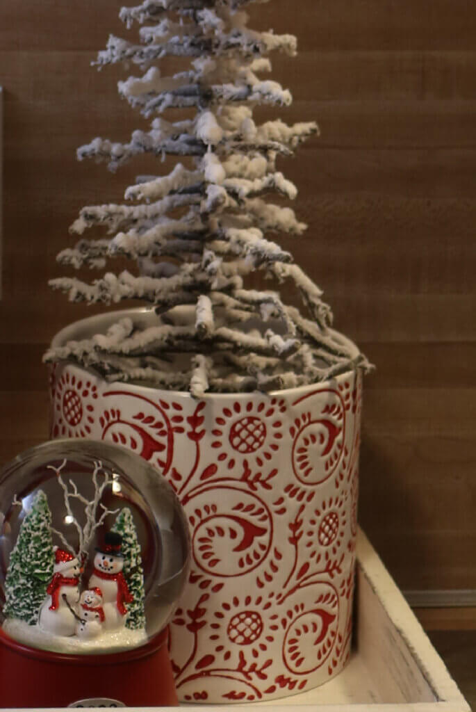 A white wood tray with a little Christmas tree inside a red and white plant pot and a Christmas snow globe.