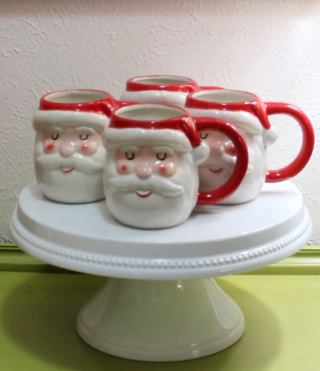 In the Christmas kitchen, there is a white plate stand, with beading textured detail, sitting on my coffee bar holding Santa mugs.