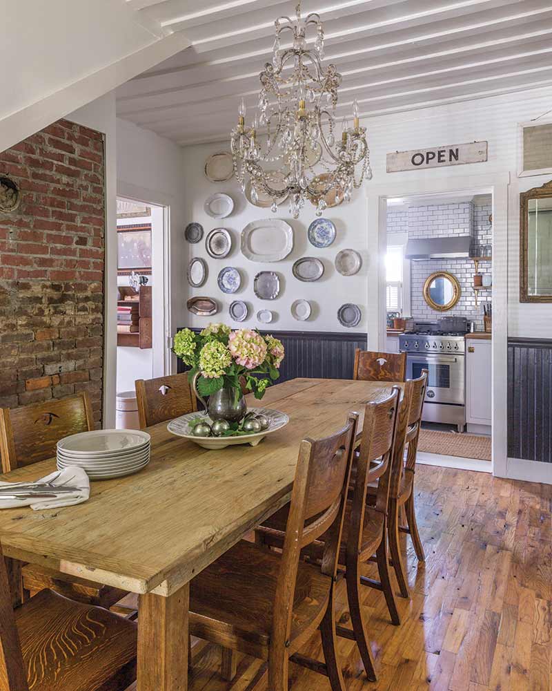 In A Charming Cottage In Tennessee, the dining space has a rustic wood table and chairs with a brick accent wall.
