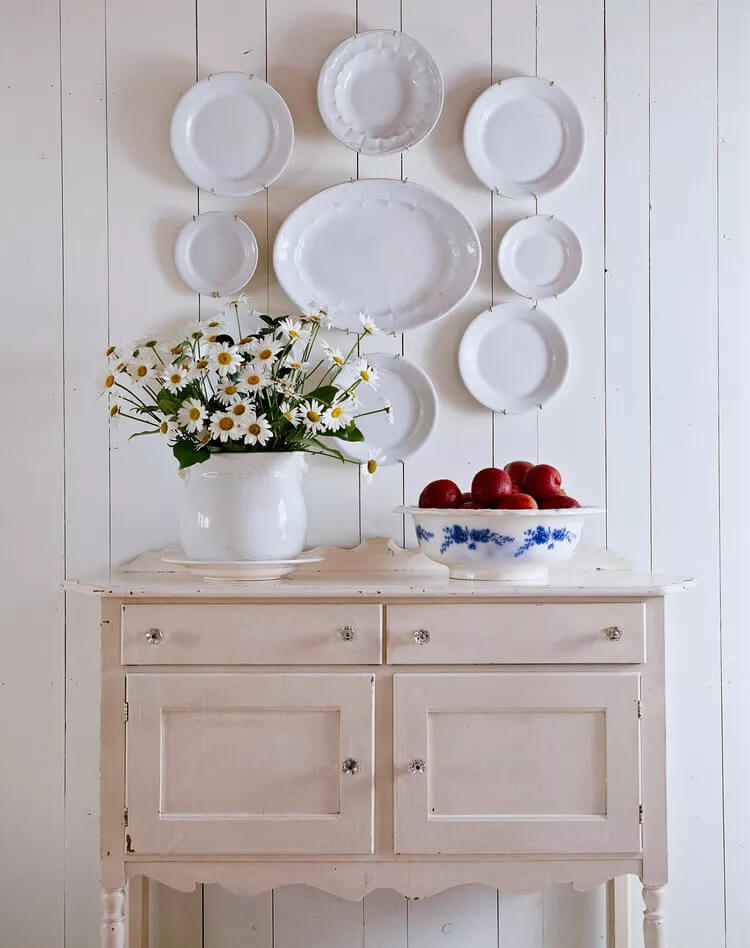Old cupboard under a well of white plates. White container of daisies and blue and white bowl of red apples.