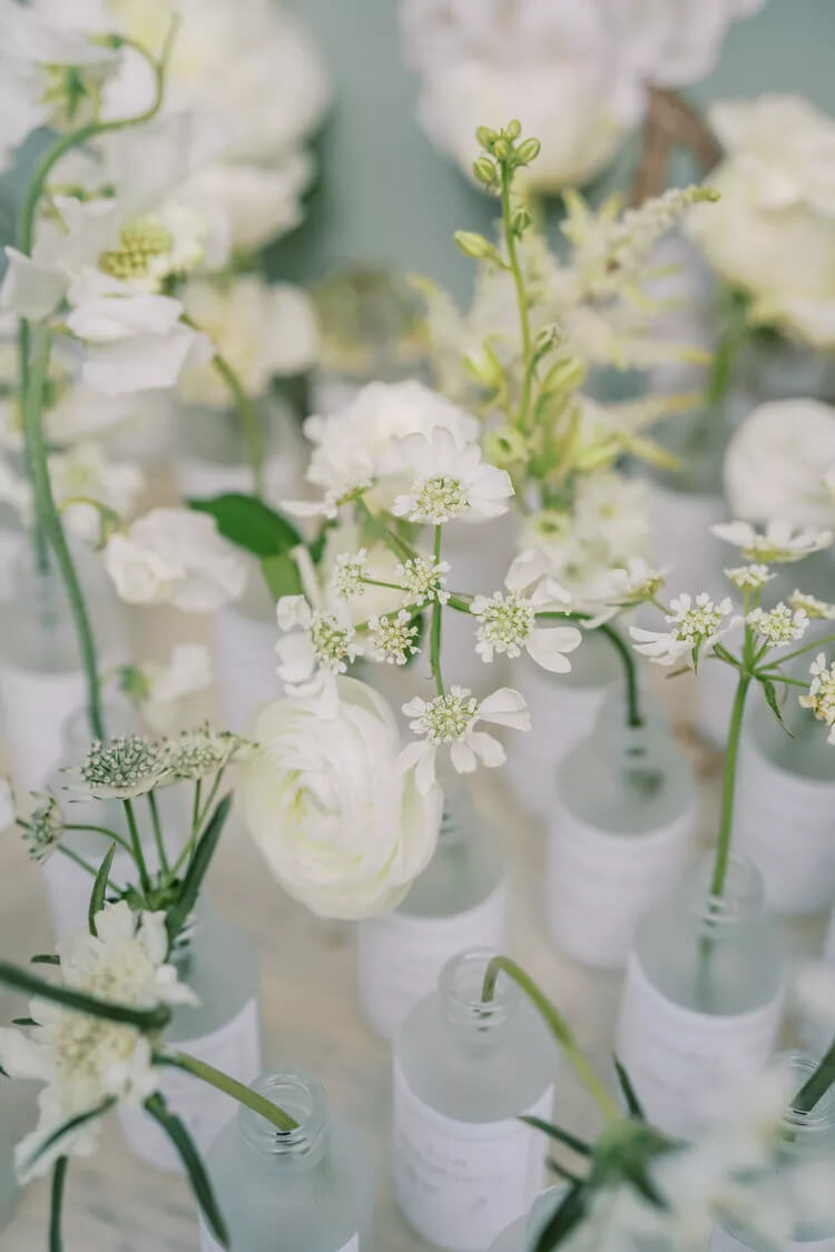 In Decorating With Vintage Bud Vases, these bud vases are holding white flowers with wedding guests seating assignments written on the side.