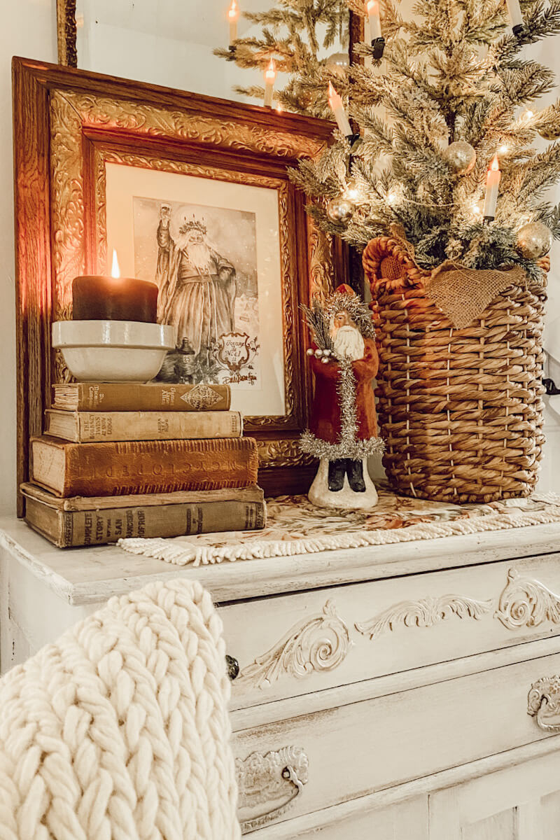A neutral display with a tree, a Santa painting, books, a Santa, and a candle in a vintage bowl.