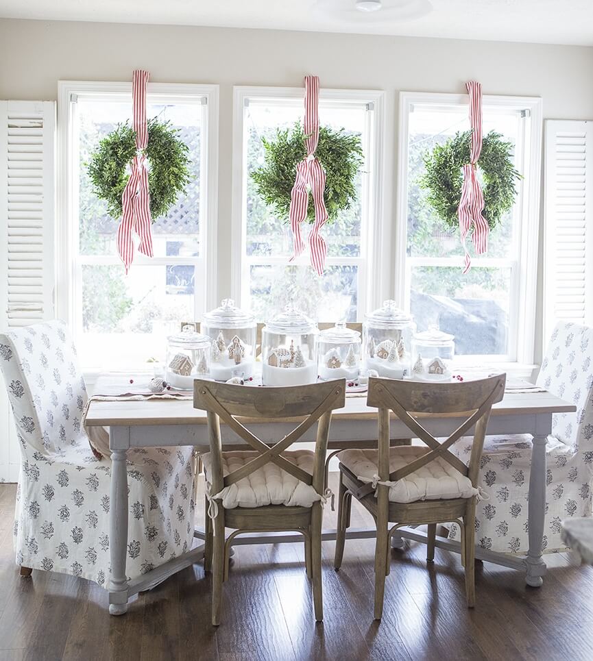 Simple green wreaths hanging at the window from red and white ribbon. Gingerbread in jars on the table.