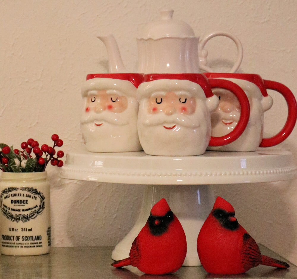 In Dining Room Santa Vignettes, Santa mugs on a cake stand with two male cardinals underneath