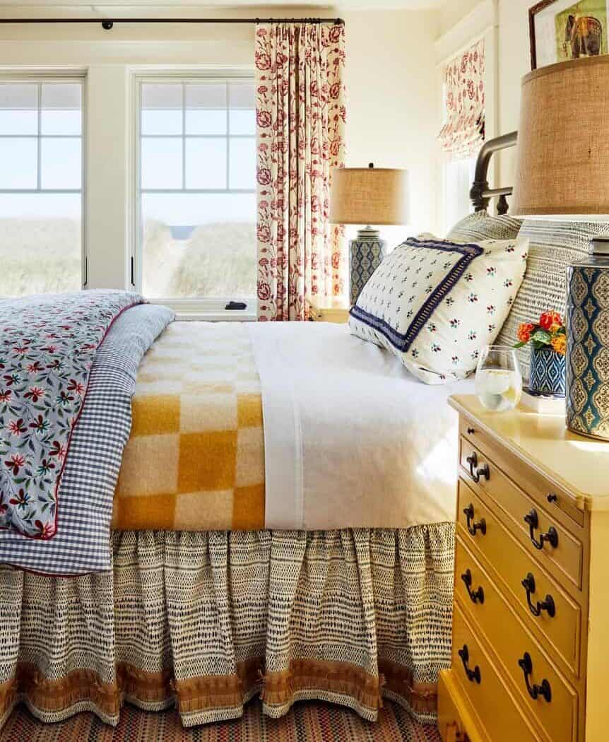 In The Love Of Home Files #18, this is a bedroom with colorful linens on the bed, two blue patterned lamps, and red and cream curtains on the window.