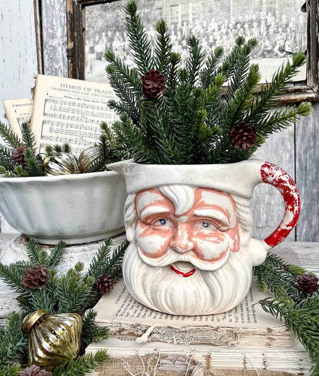 A chippy mug filled with green sprigs next to a white bowl with sprigs, gold ornaments, and sheet music. A vintage framed picture is above it on the wall.