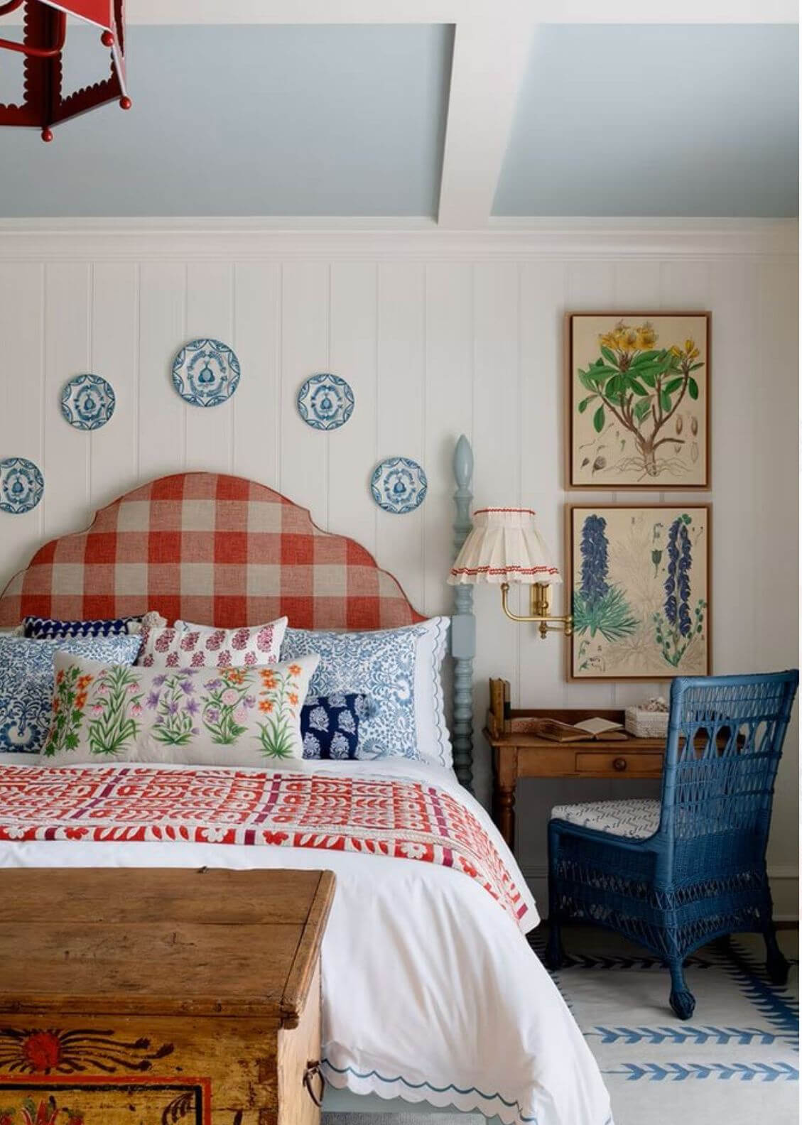 Red, white, and blue bedroom with upholstered headboard, various pillows, and a wooden desk with blue wicker chair next to bed.
