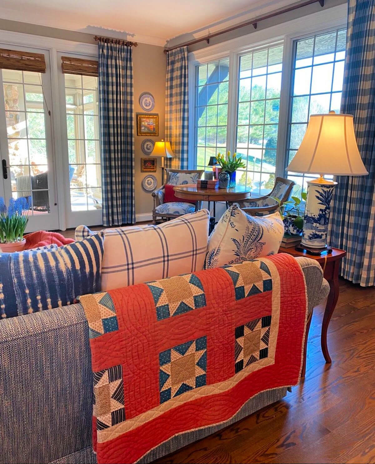Red, white, and blue living room with a quilt over the back of the couch, blue and white checked curtains, and two chairs with table in front of the window.