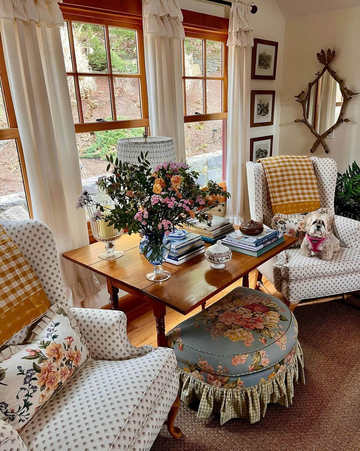 Grannycore style room with two upholstered chairs, quilts, and a table in the middle in front of windows.