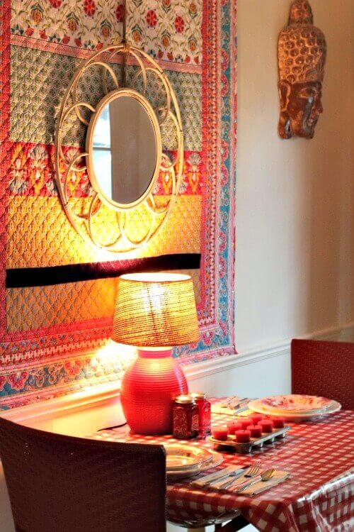 An old dining space with a boho quilt on the wall with a mirror hanging on it. The rectangular table has a red and white checked oilcloth, a red lamp, and red and white floral dishes and candle in a muffin tin. Red salt and pepper shakers and red chairs.