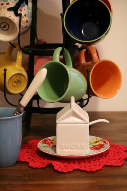 A metal mug holder with different colored ceramic mugs. A white sugar container on a floral dish.