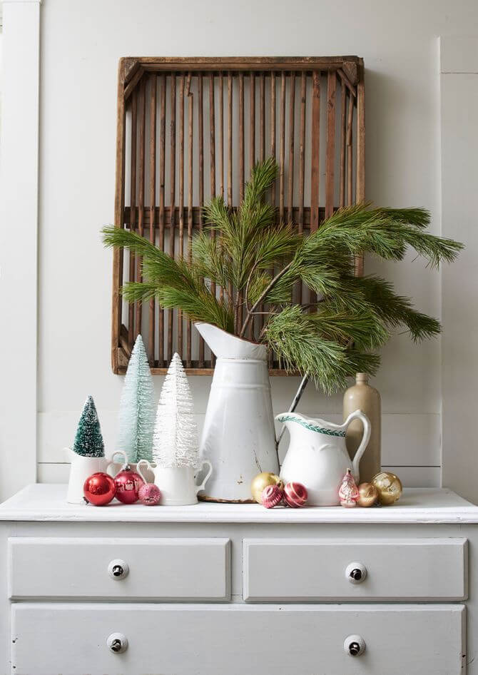 White pitchers on a white dresser holding greenery and bottle brush trees. Scattered ornaments around them and a wooden basket overhead.