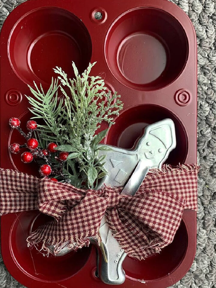 Budget-Friendly DIY holiday decorations is this red painted muffin tin with greenery and red berries, plus a gingerbread man cookie cutter tied with torn gingham fabric strip.