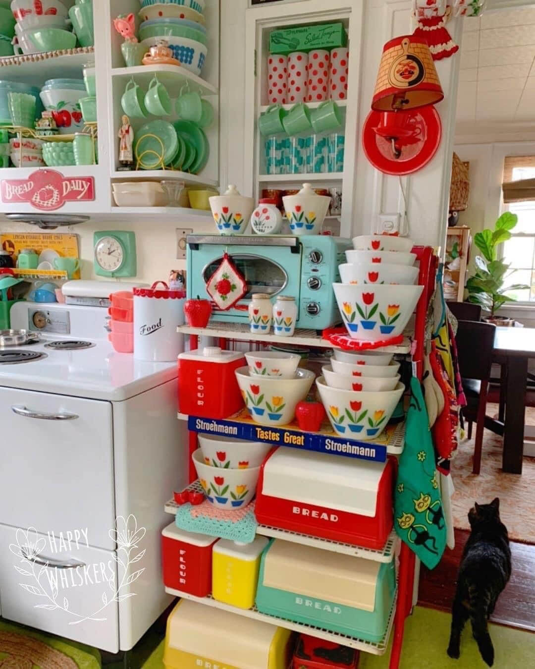 A fully retro kitchen with a collection of vintage bread boxes and Pyrex, plus Jadeite dishware and other colorful decor above. A black cat is walking from the room. Could be Simon!