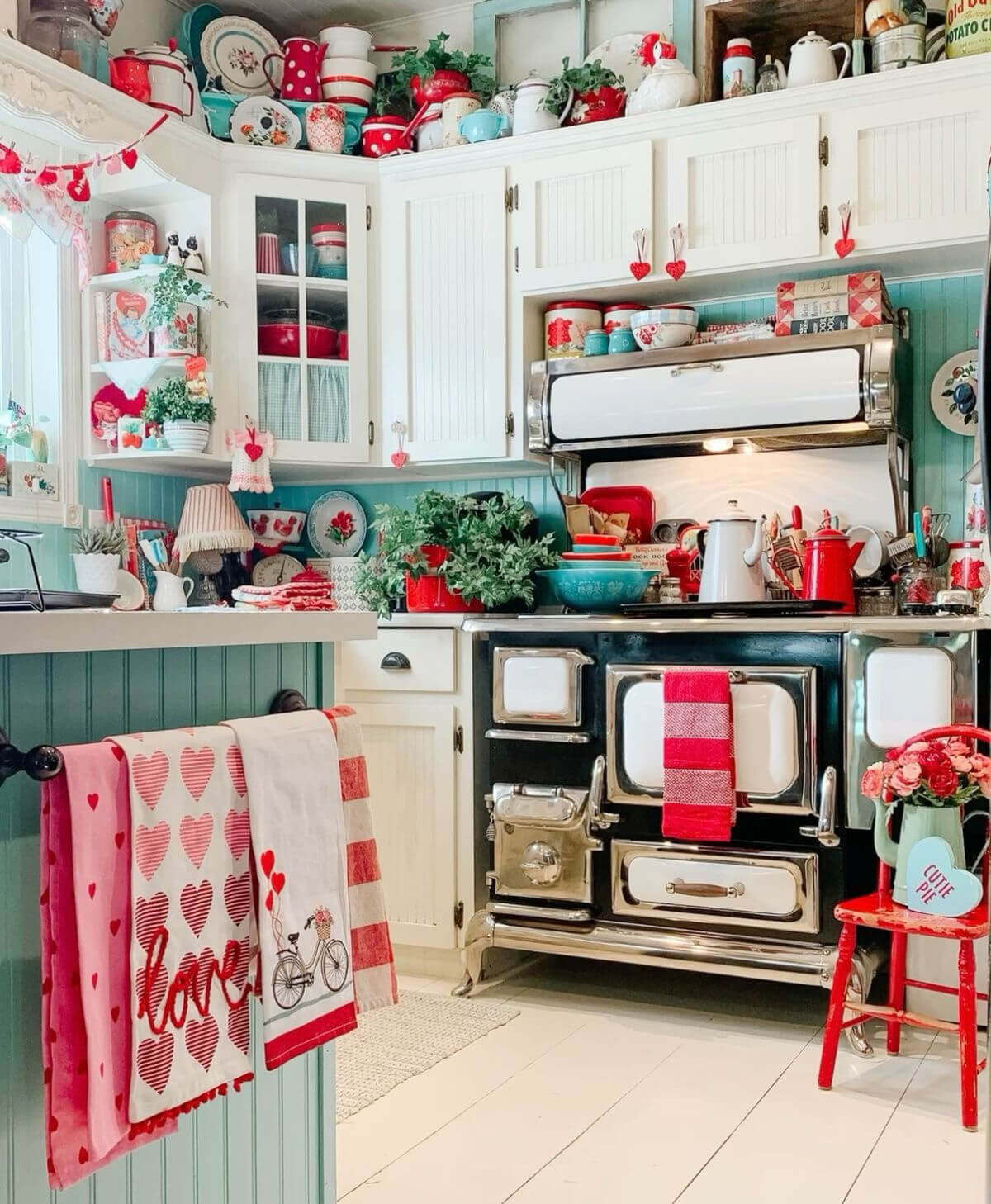 A red, white, turquoise, and black kitschy retro with an old stove, and a red chair off to the side. Fun red, white, and pink dish towels hang on the side of the cabinets. On the countertop are lots of fun details that befit a kitchen from that time.