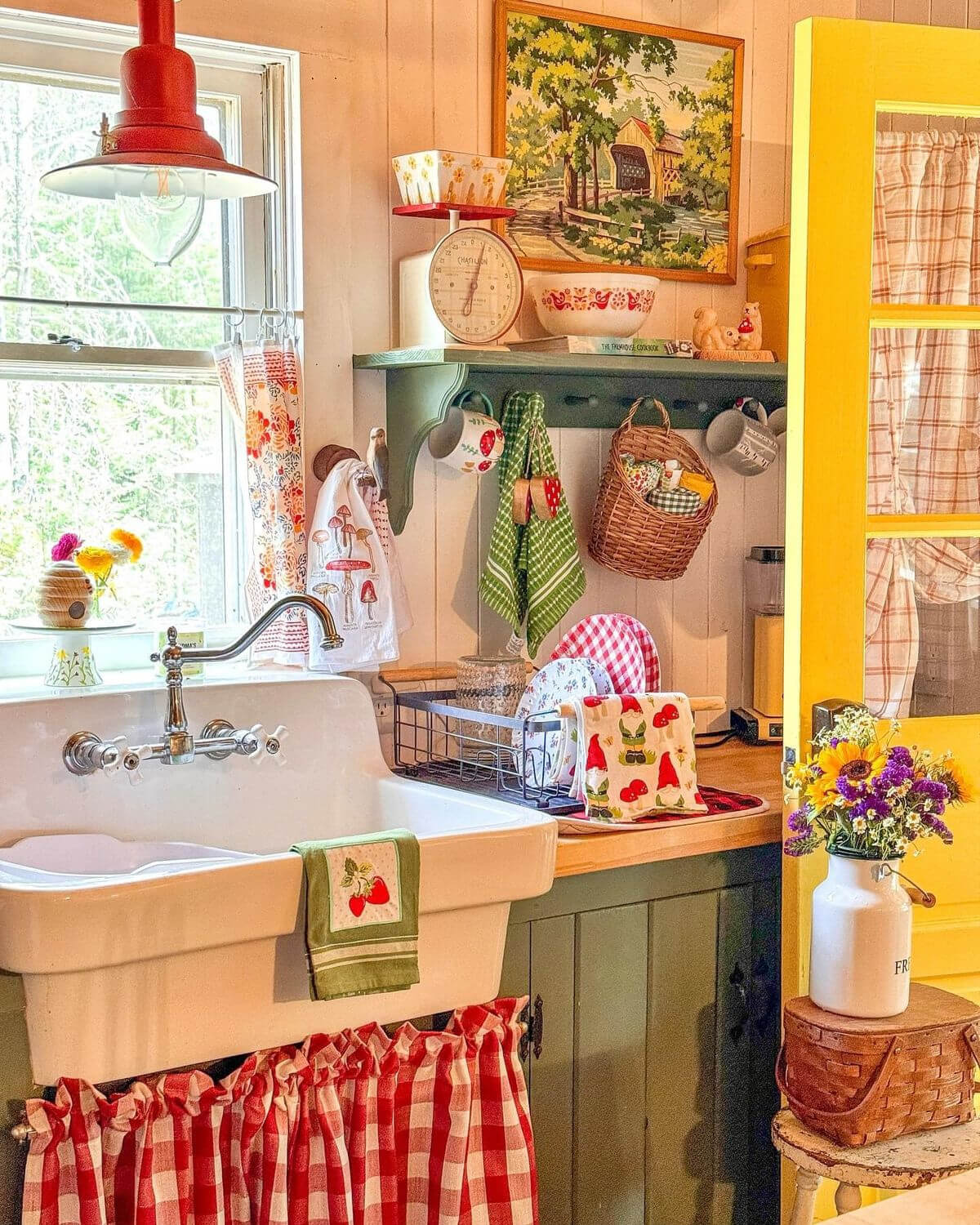 A cheerful, kitschy kitchen, a white farmhouse-style sink, and a red-and-white-checked curtain beneath. On the wall is a green shelf filled with vintage decor and linens.