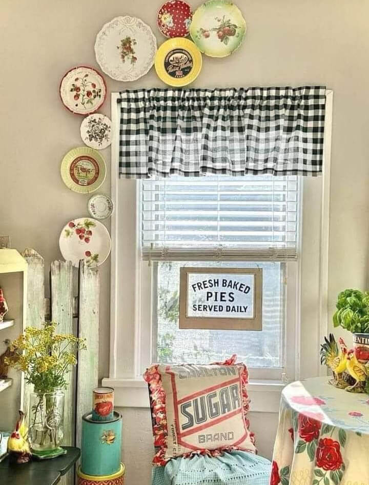 A kitchen with a round table covered with red roses, and a framed sign that says fresh-baked pies served daily. Then there's a black-and-white checked valance, dishes hanging on the wall, and a "sugar" decal pillow in the chair.
