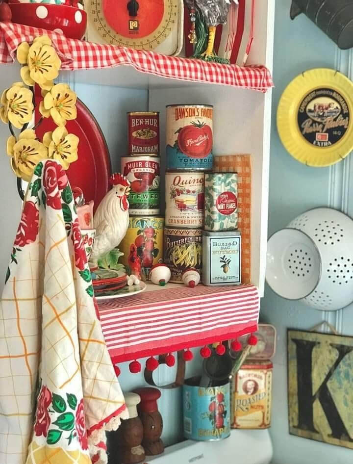 A corner of a kitschy kitchen, with a red-and-white-striped linen hanging from the upper open cabinet. On the wall hangs a white colander, a "K" sign, along with vintage-style cans in the cupboard.
