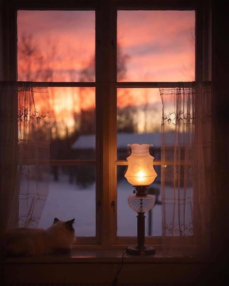 A candle stares outside a window at the snowy landscape while a light softly glows inside.