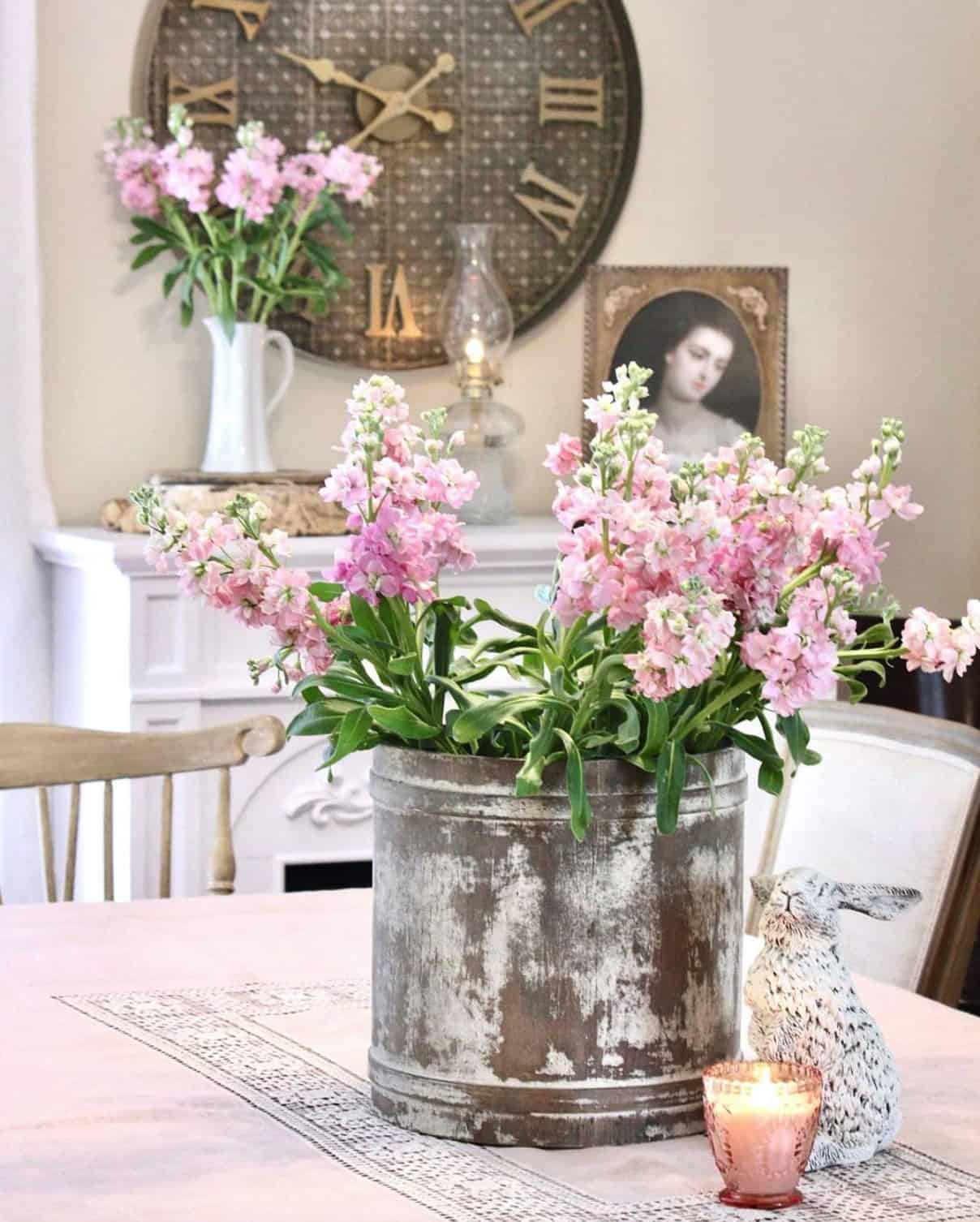 A table centerpiece of a rustic container and pink flowers. A rabbit hovers nearby with a pink candle.