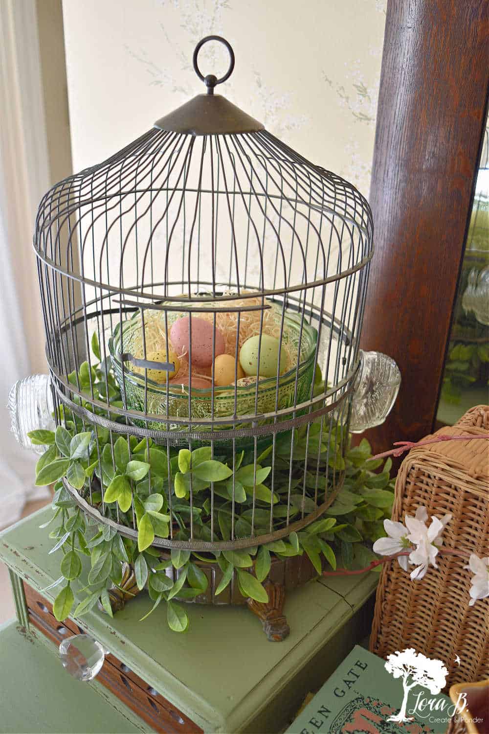A vintage bird cage holds greenery and a green glass container with pastel colored eggs.