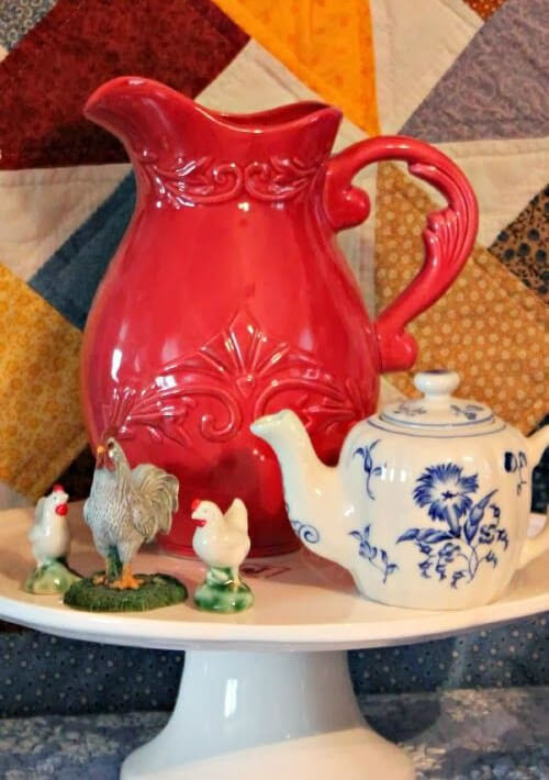 Put decorative vignettes on display with a small cupcake stand, which holds a red pitcher, a blue and white creamer, and chickens.