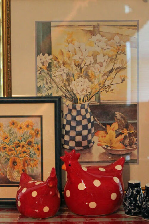 Red and white polka dotted ceramic chickens in front of layered paintings.
