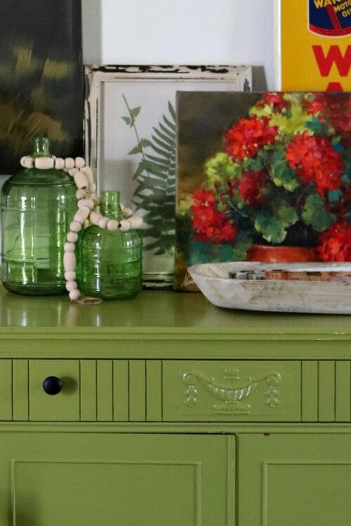 My green sideboard with green bottles and layered paintings against the wall.