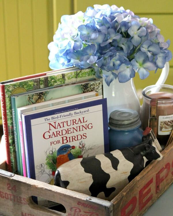 A wooden pepsi box with books, a cow, a painted vintage jar, a candle, and a white pitcher with blue hydrangeas.