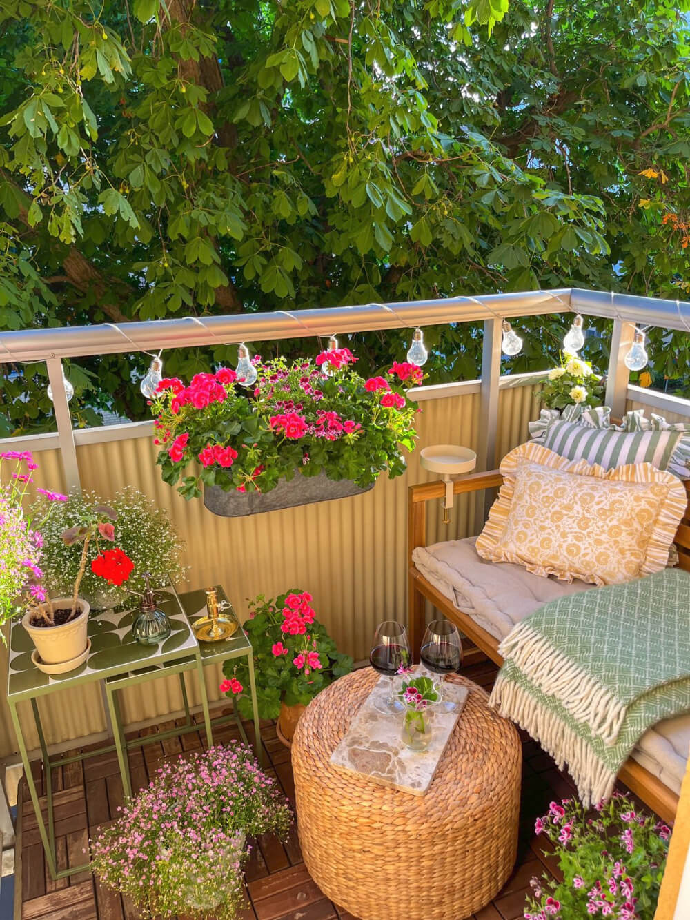 A wooden bench with pillows, round table with glasses of wine, nesting tables with plants, and geraniums hanging on the railing.