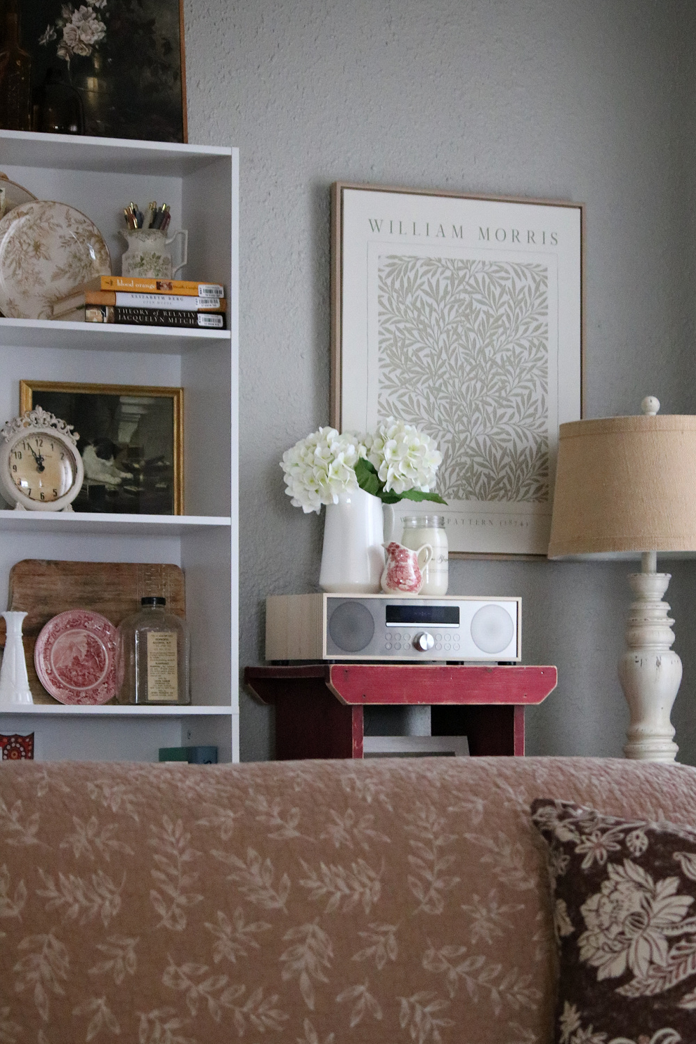 A corner of my living room with my bookshelves, a cupboard with drawers, a lamp, my CD player, and faux hydrangeas in a pitcher, a candle, and a transferware creamer. Over this is a William Morris print.