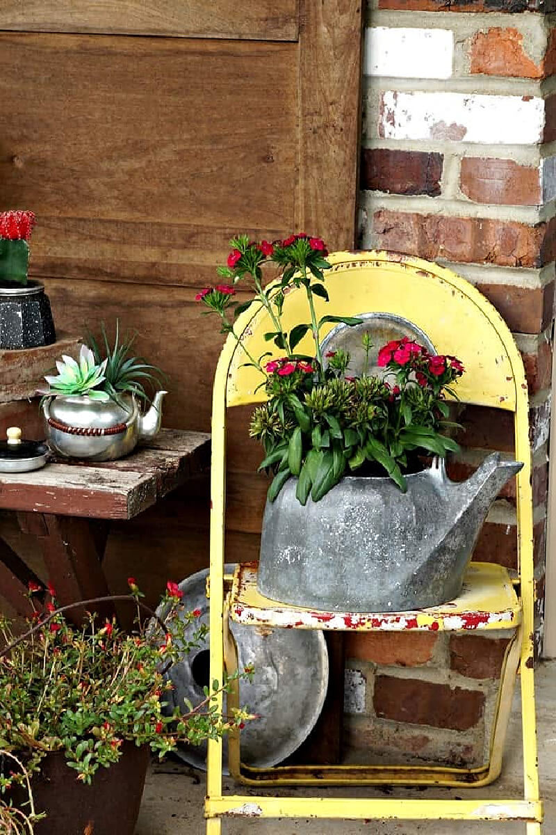 Repurposed treasures for a cottage garden worn tea kettle with flowers planted inside sitting on a yellow chair.