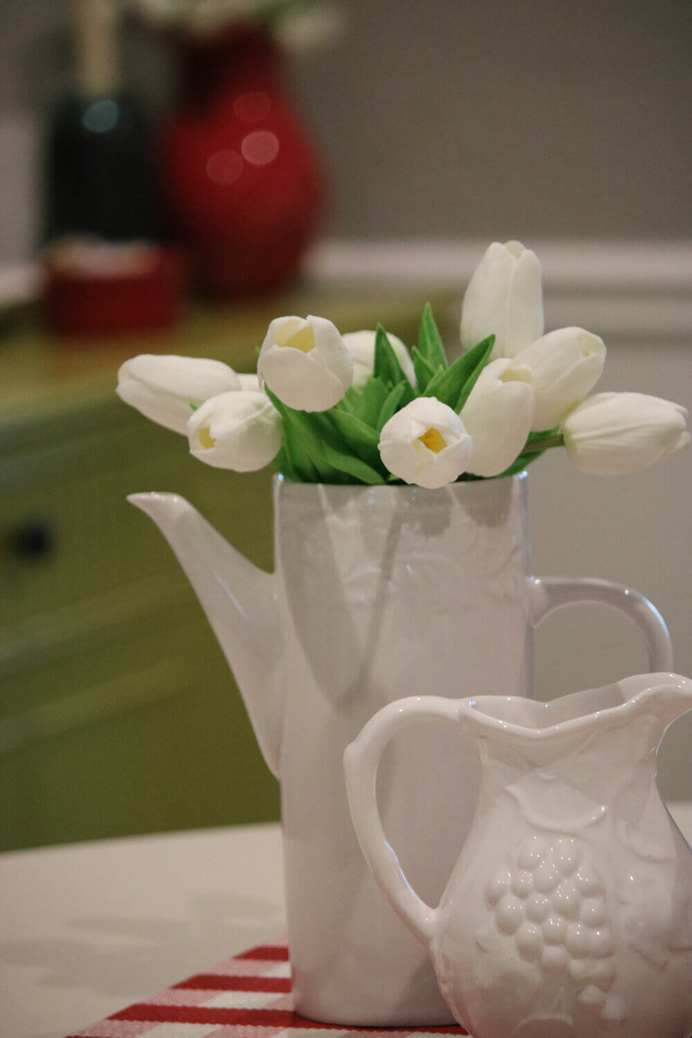 A different centerpiece of simple white tulips in a white pitcher next to another smaller white pitcher on a red and white checked placemat.