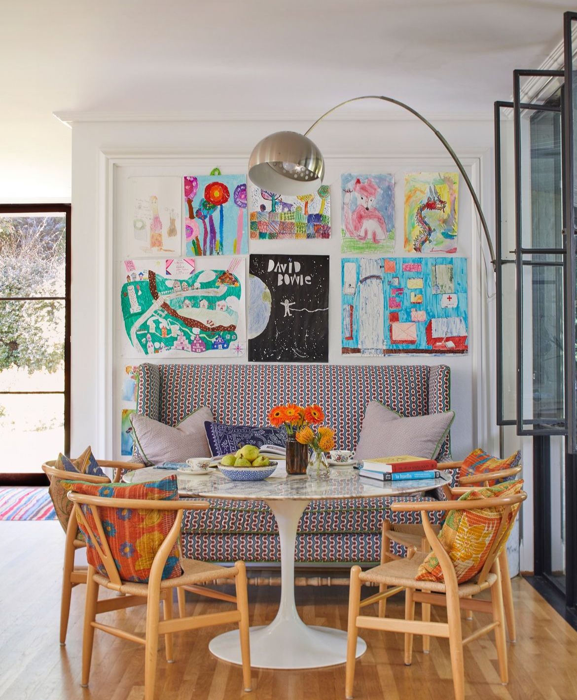 A corner with a patterned loveseat, a round tulip tip table, and four wooden chairs. Lots of amateur art above the loveseat.