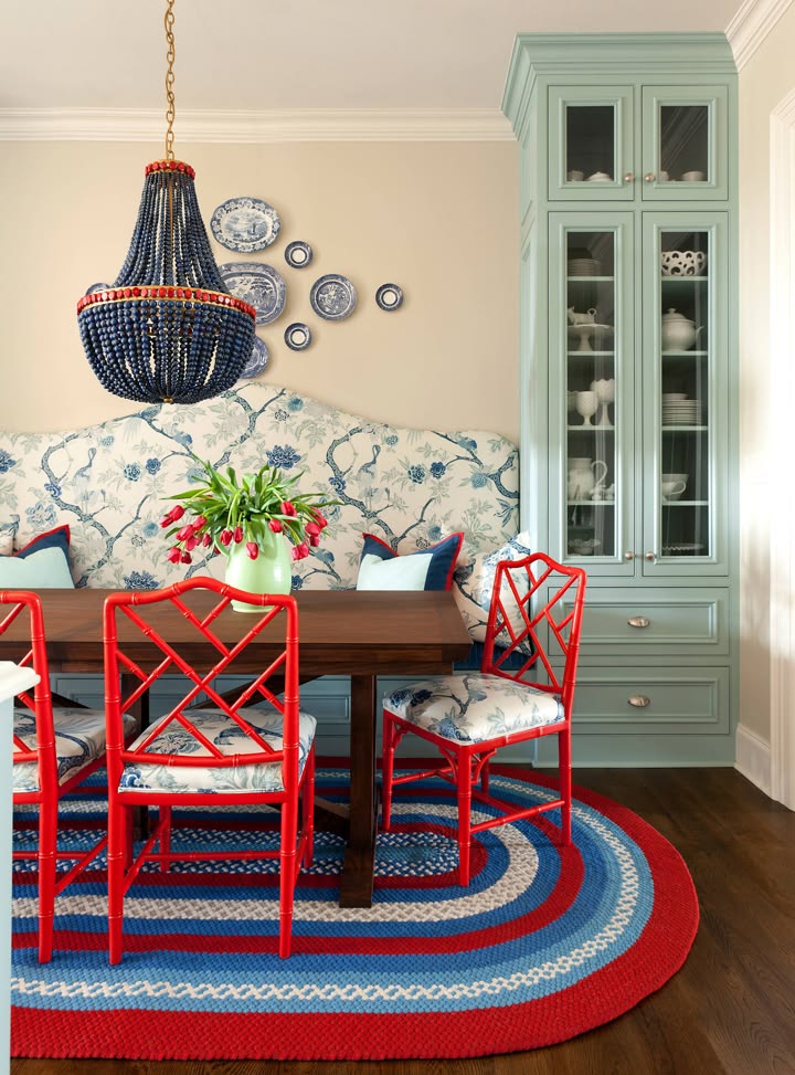 How to create a cozy breakfast nook with a blue and cream upholstered bench, a brown wooden rectangular table, and red chairs with the same upholstered cushions as the loveseat. A red, white, and blue braided rug grounds the room.