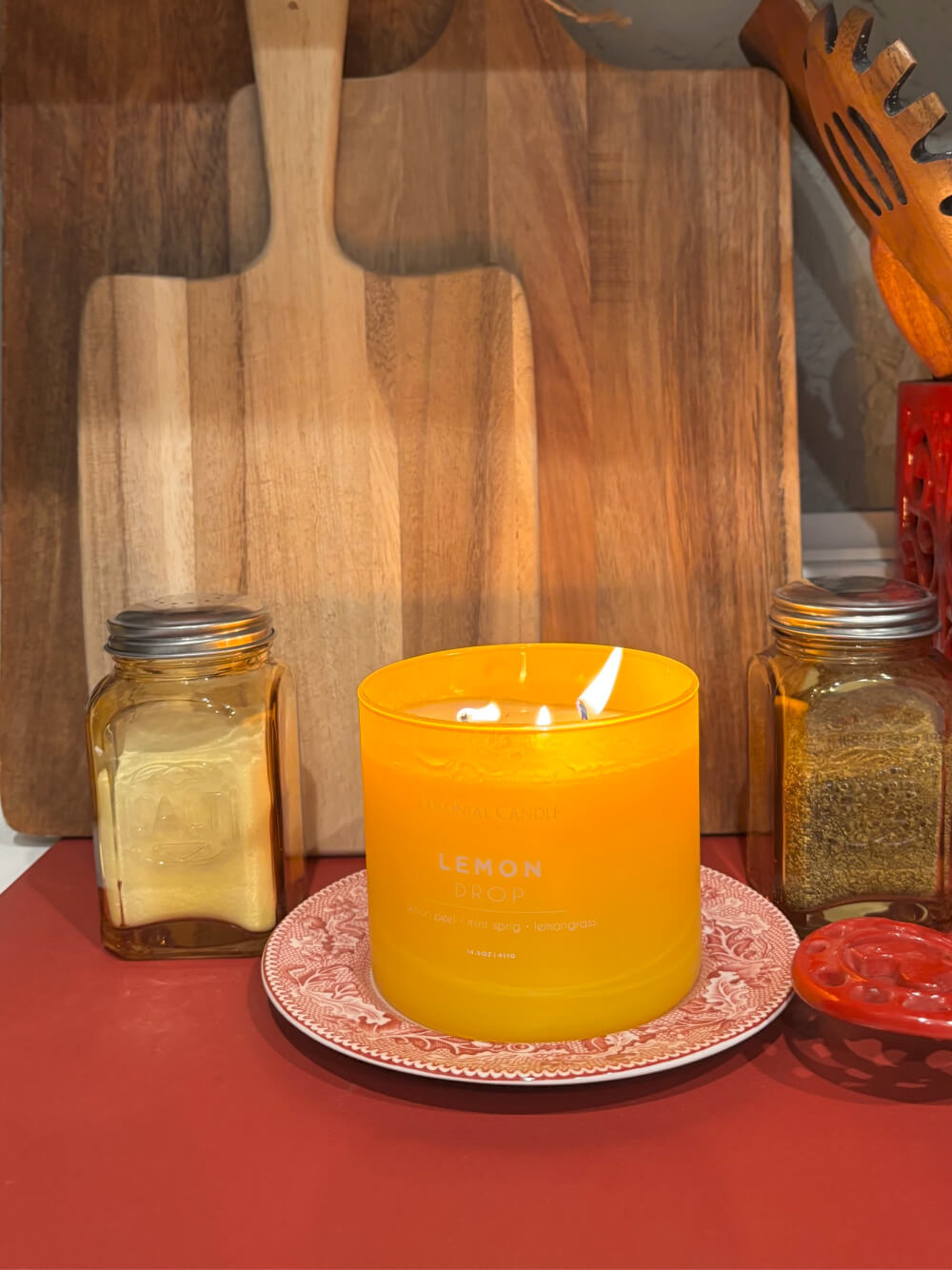 A lemon drop candle on a red and white transferware dessert dish. Gold salt and pepper shakers surround it in front of a collection of cutting boards.