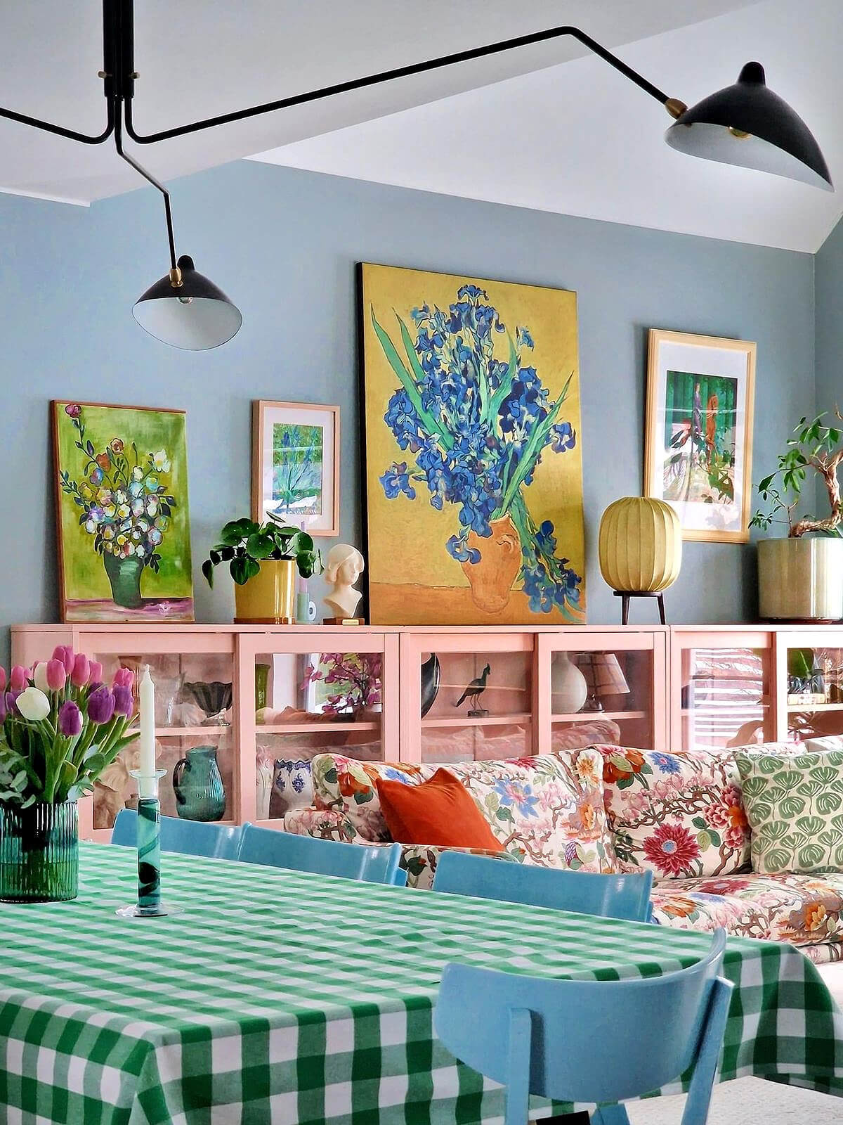 A floral couch with pink glass-fronted shelves behind it. A long rectangular dining table with green and white tablecloth and blue chairs.