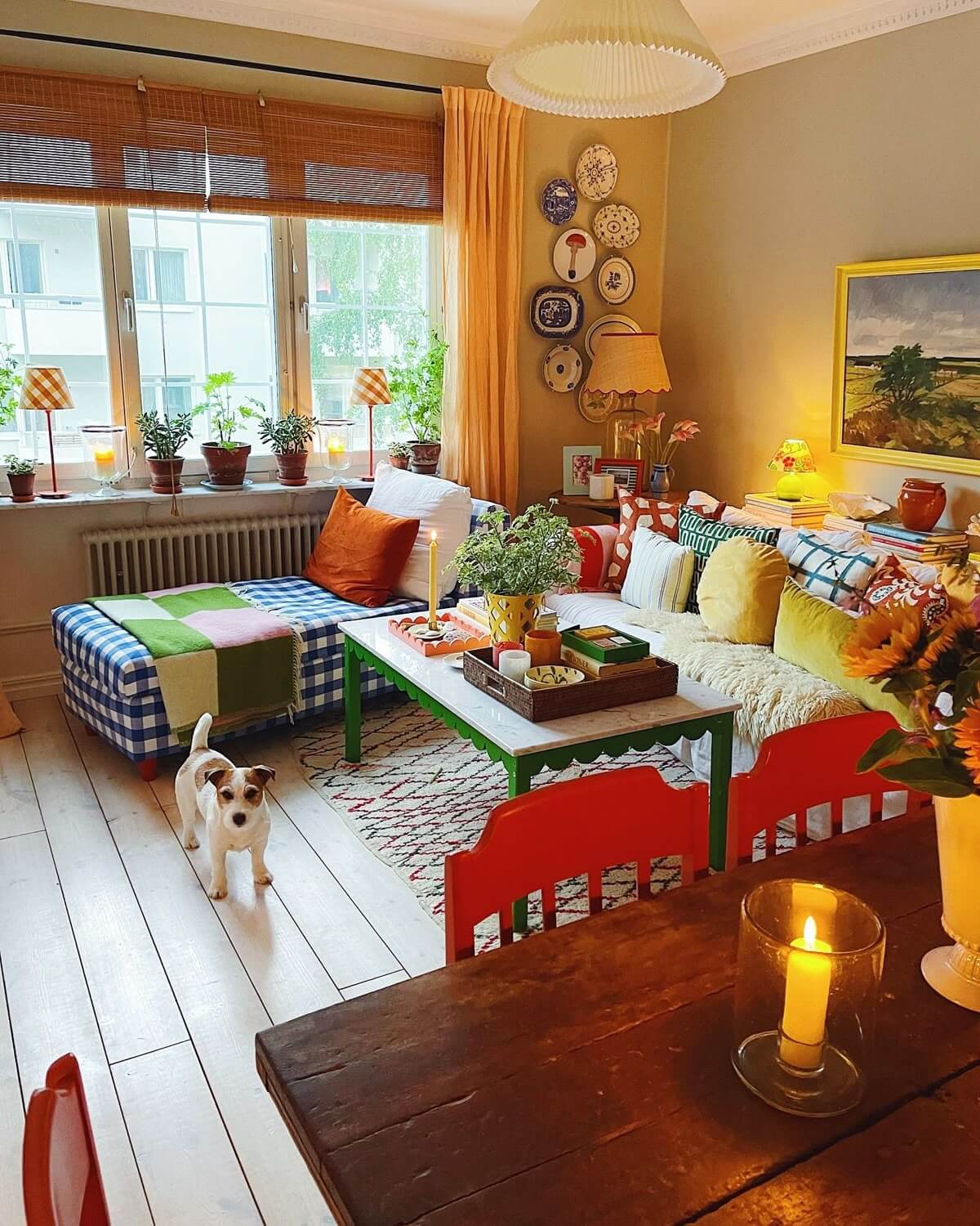 This colorful space has a couch and a blue and white checked chaise. There's a marble topped coffee table with green scalloped wood embellishing it. A table with red chairs and a small dog.
