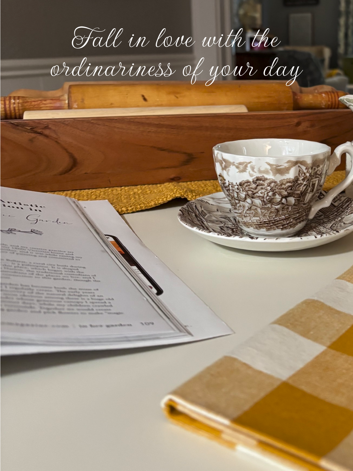 An ordinary day in my dining room with the round white table, wooden dough bowl of rolling pins. A magazine and a brown and white transferware cup and saucer.