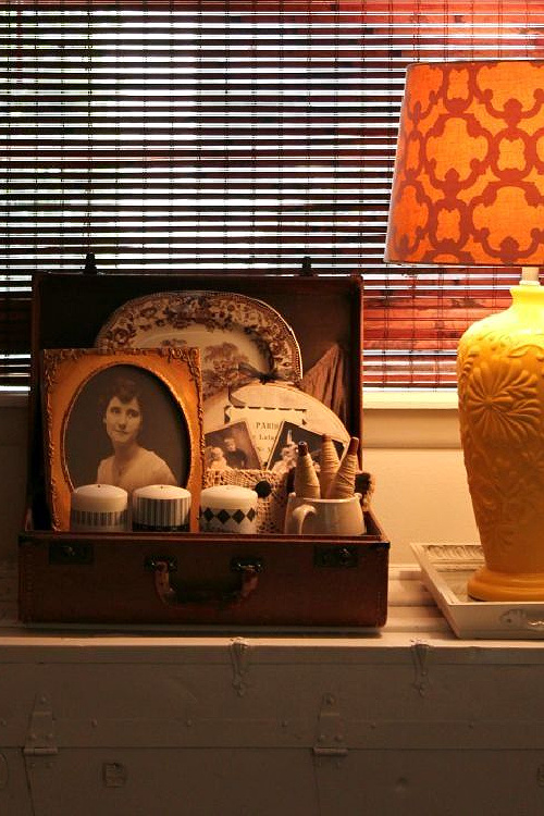 22 ideas to repurpose vintage suitcases includes this vignette on top of a white trunk in my home next to a yellow lamp.