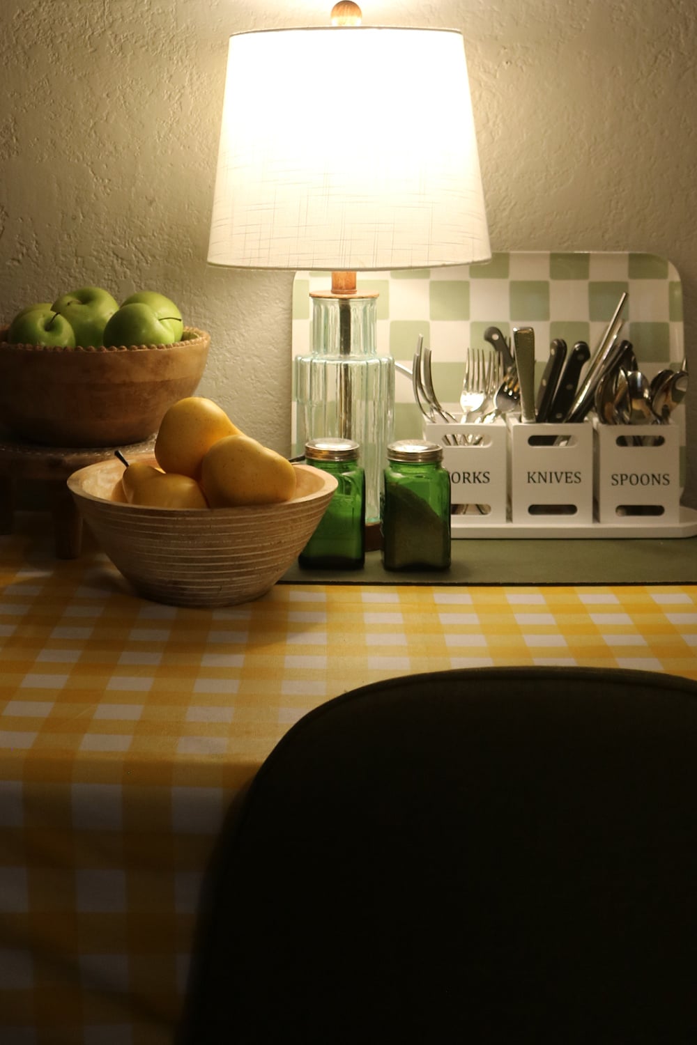 In how to create a cozy afternoon tea, this is a table with a yellow and white checked tablecloth, a bowl of pears, green salt and pepper shakers, and knives, forks, and spoons.