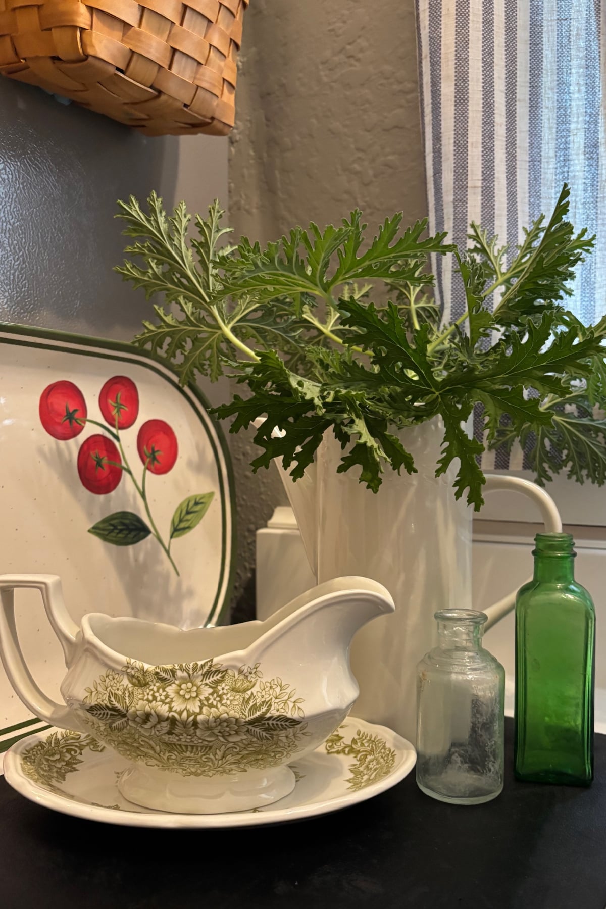 A white pitcher of geranium leaves, a platter with tomatoes, two vintage bottles, and a green and white gravy boat.